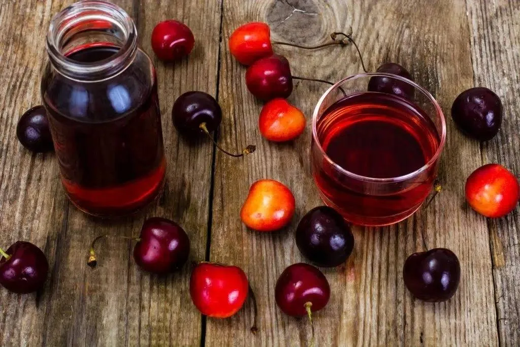 A glass and jar of cherry juice on a rustic wooden surface, surrounded by fresh cherries.