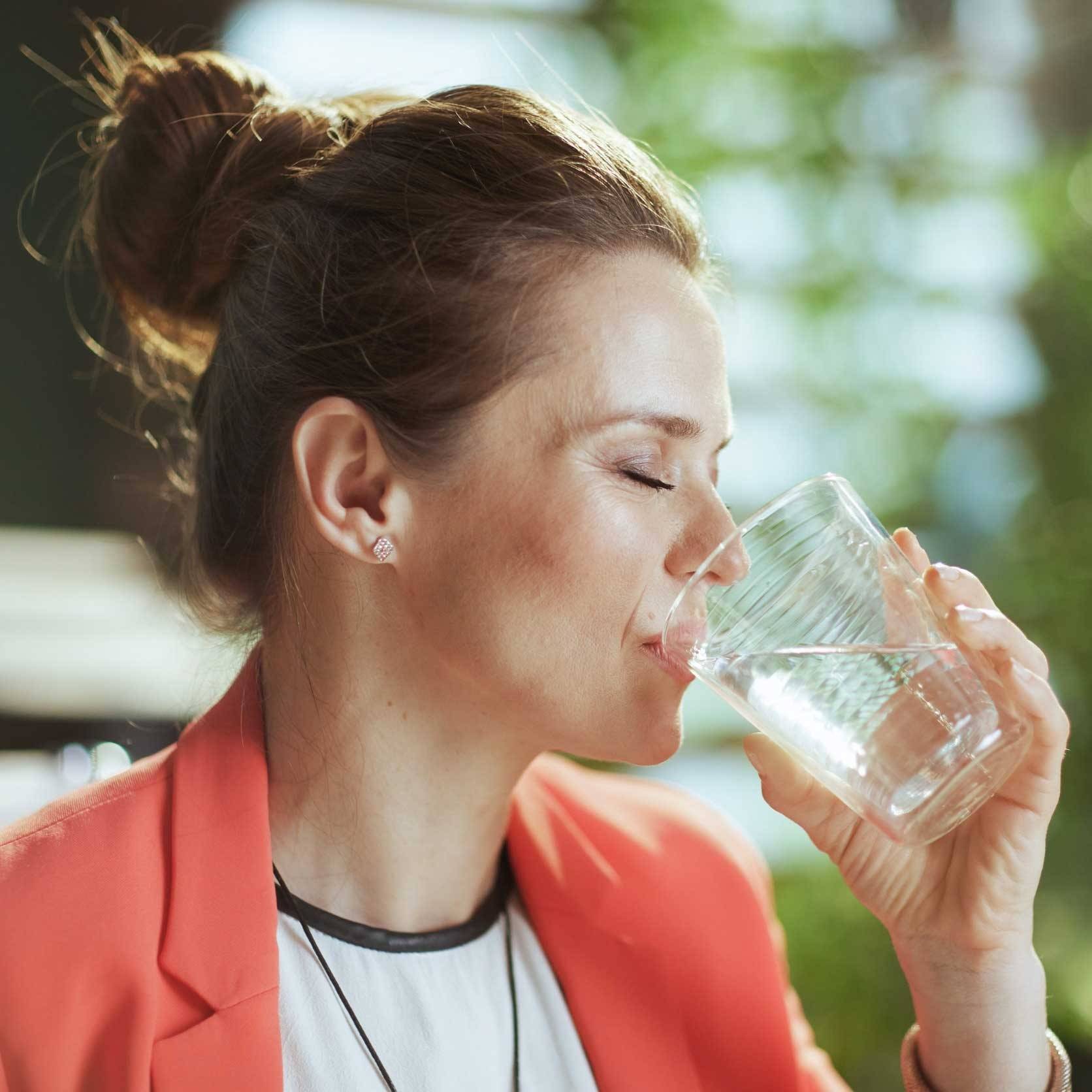 Woman with brown hair in a bun drinks water, wearing a coral blazer and white top; blurred green background.