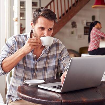 Man in plaid shirt sits at a table with a cup and laptop, browsing pain relief supplements; another person is behind.
