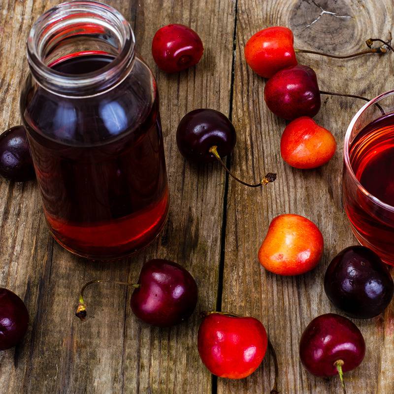 A glass jar and a glass of red juice sit on a wooden table with cherries scattered around, evoking freshness.