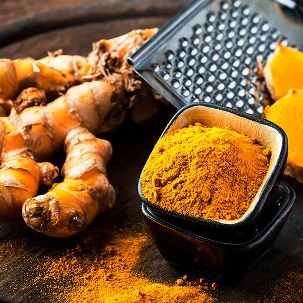 A black bowl of turmeric powder sits beside fresh turmeric roots and a grater on a dark wooden surface.