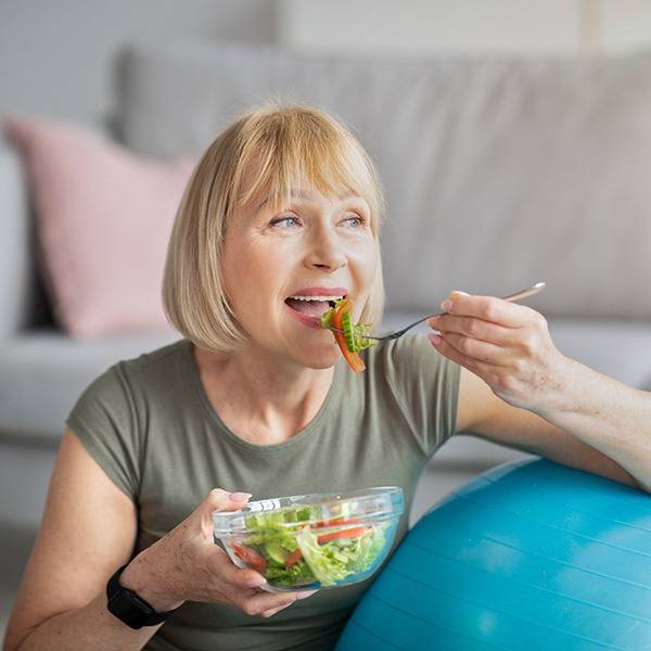 Older woman with short blonde hair eats salad next to a blue exercise ball, emphasizing wellness and supplements.