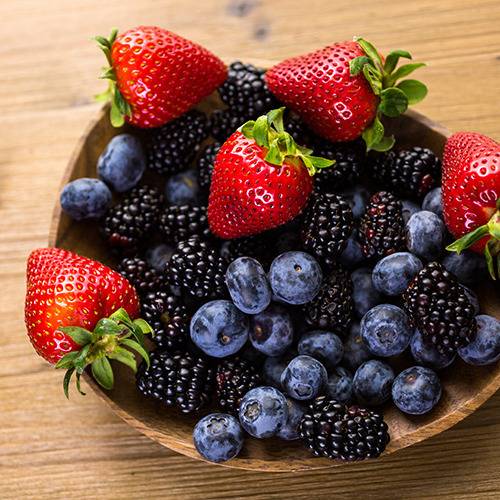 A wooden bowl of fresh strawberries, blueberries, and blackberries sits next to natural supplements on wood.
