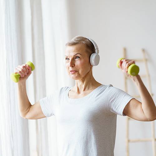 Older woman in headphones lifts small green dumbbells indoors, exercising and staying focused with supplements nearby.