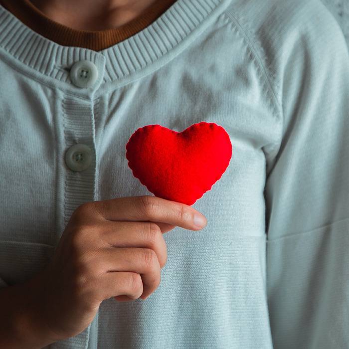 Person in a light blue sweater holds a small red fabric heart, symbolizing care and wellness.
