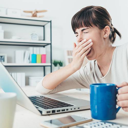 A woman at a desk with a laptop yawns, covering her mouth as she reaches for her blue mug of supplements.