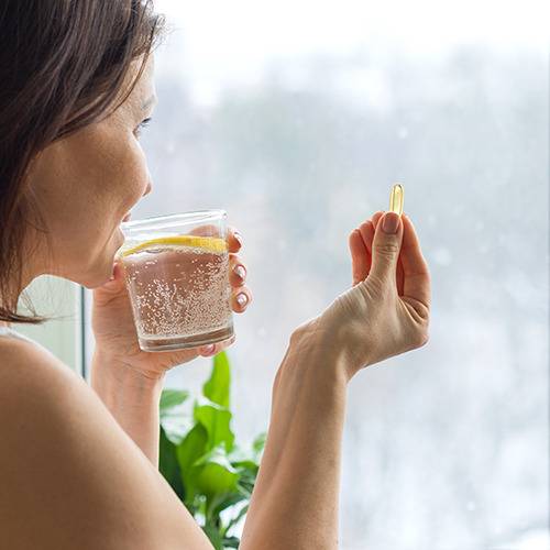 A woman holds a glass of lemon water and a yellow capsule, looking thoughtfully out the window.