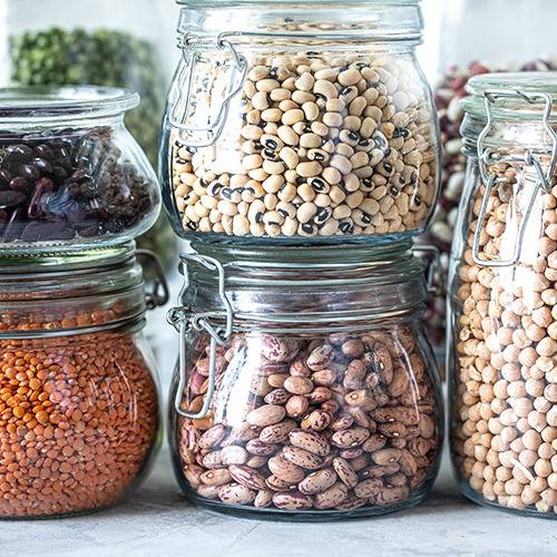 Five glass jars with assorted dried beans and lentils are grouped together on a light surface.