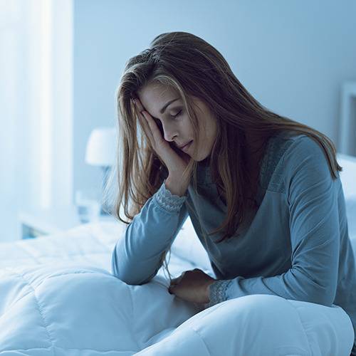 A tired woman in pajamas sits on her bed in a dimly lit room, possibly thinking about natural pain relief.