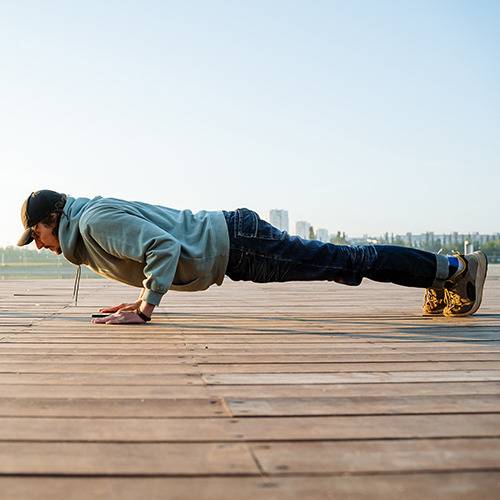 A person in jeans, hoodie, cap, and boots does a push-up outside with a city skyline in the background.