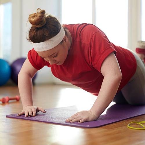 A person in a red shirt does knee push-ups on a purple mat in a bright room with natural supplements and exercise gear.