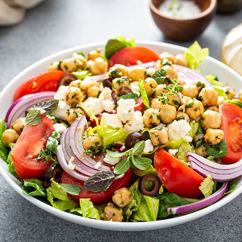 A bowl of salad with chickpeas, tomato, red onion, olives, lettuce, feta, and herbs. Supplements nearby.