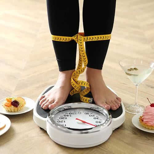 Person on a scale with measuring tape, desserts, and supplements scattered on the floor.