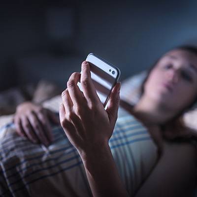 Person in bed under a plaid blanket, looking at a smartphone about natural sleep supplements in dim light.