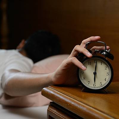 A person in bed reaches to turn off an alarm clock; a bottle of supplements sits on the wooden nightstand.