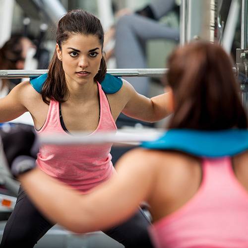 A woman in a pink tank top checks her squat form with a barbell, motivated by surgeon-formulated supplements.