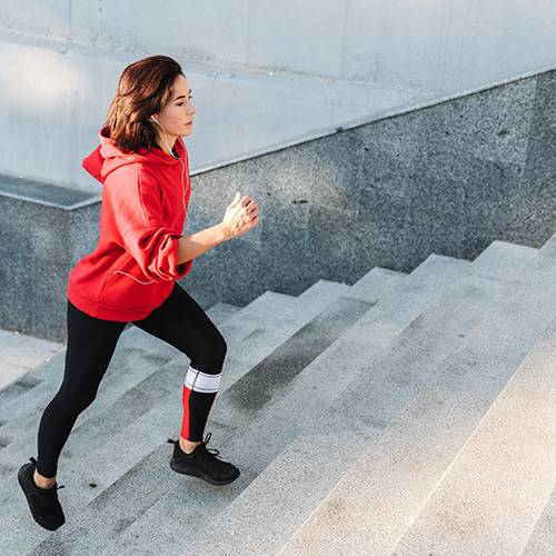 Woman in a red hoodie and black leggings runs up stairs, supported by natural surgeon-formulated supplements.