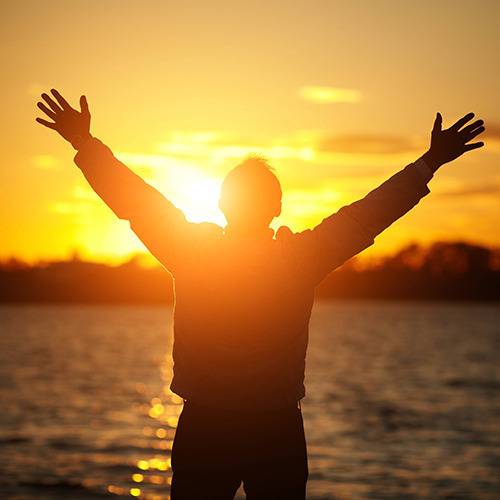 A person stands by the water at sunset with arms raised, silhouetted against the sun, embodying natural vitality.