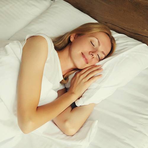 A smiling blonde woman lies in bed with her eyes closed after taking natural pain relief supplements.
