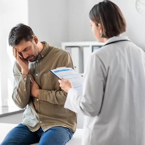 A man sits on an exam table holding his head and chest as a doctor reviews a form for pain relief supplements.