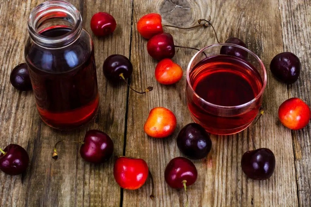 A glass and a jar of cherry juice sit on wood, surrounded by scattered red and dark cherries.