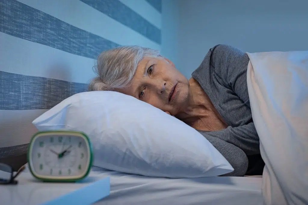 An older woman lies in bed under a white blanket at 3:09 AM, possibly seeking natural ways to sleep better.
