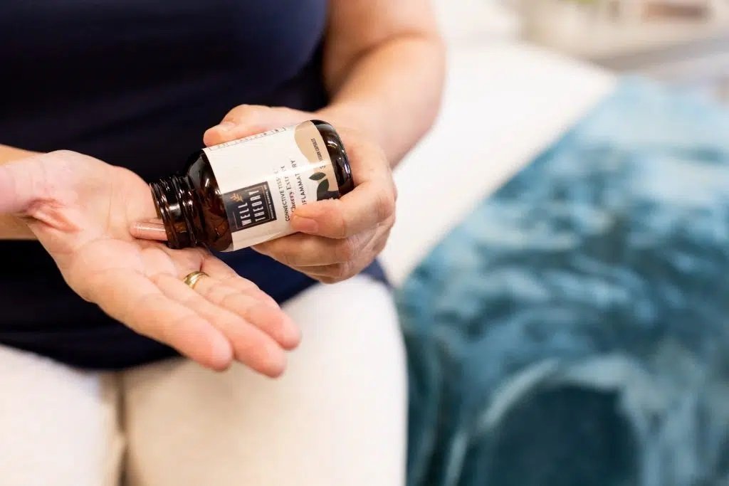 A person pours natural supplements from a brown bottle into their hand while sitting on a bed with a blue blanket.