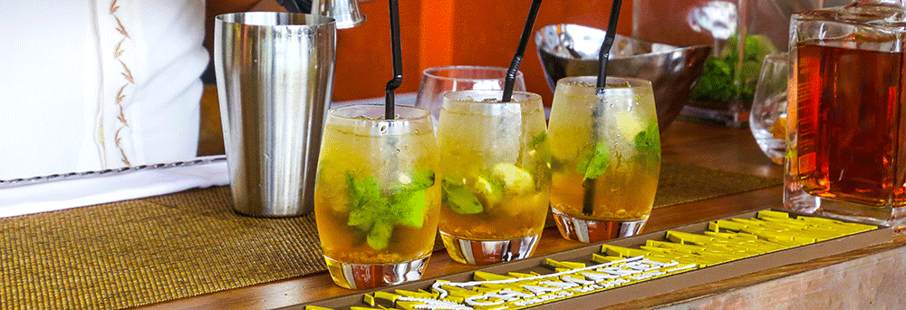 Three mojito cocktails with mint and lime sit on a bar counter beside natural supplements and a metal shaker.