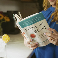 Person in blue shirt reads "Bone on Bone" next to a glass with lemon water, exploring supplements.