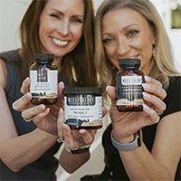 Two women smiling, holding bottles and a jar of supplements toward the camera.