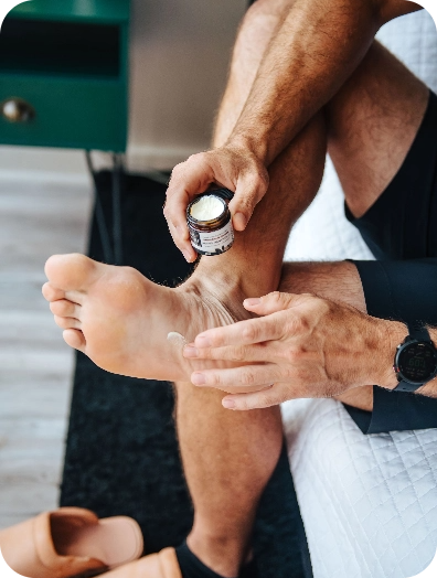 Sitting on a bed, a person applies cream to their foot; pain relief supplements and sandals are nearby.