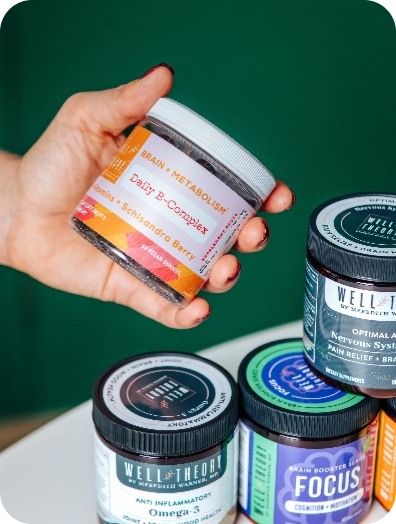 A hand holds a "Daily B-Complex" jar above stacked Well Theory surgeon-formulated supplement jars.