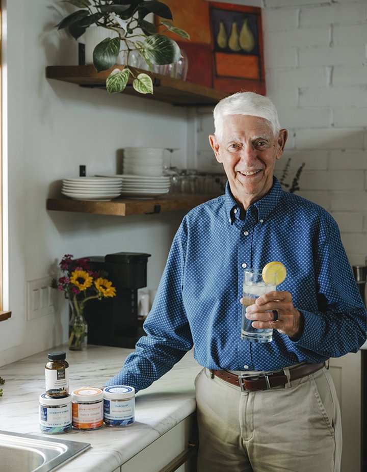 An older man in a blue shirt stands in a kitchen holding a lemon drink; supplements and cans are on the counter.