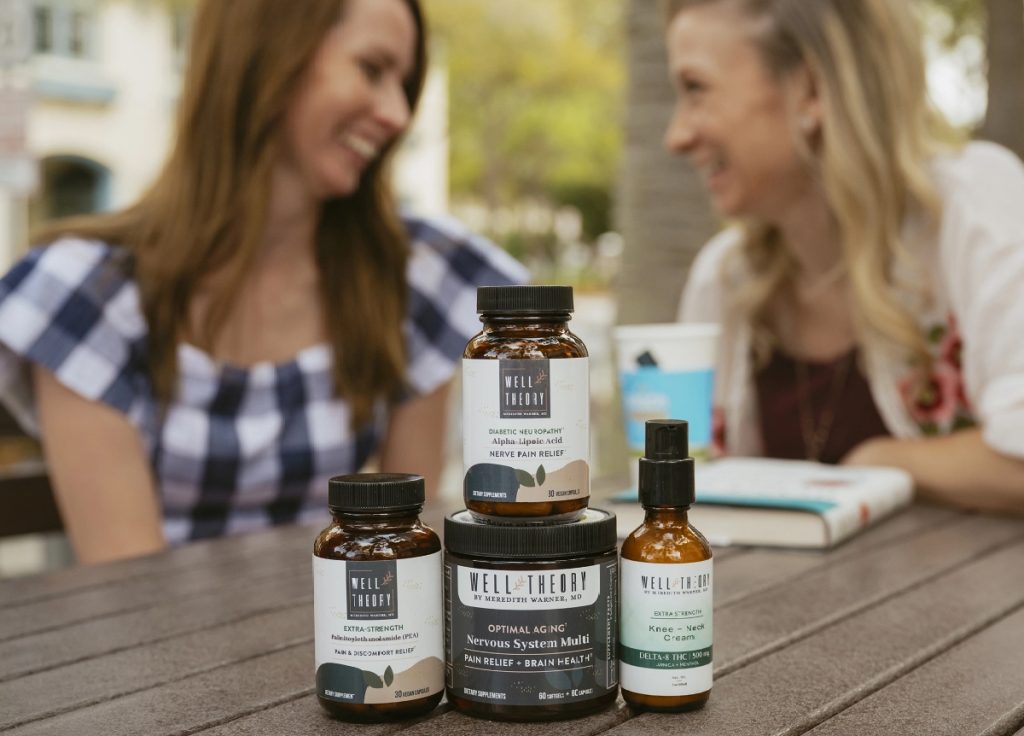 Two women smile outdoors with Well Theory supplements in focus on the table before them.