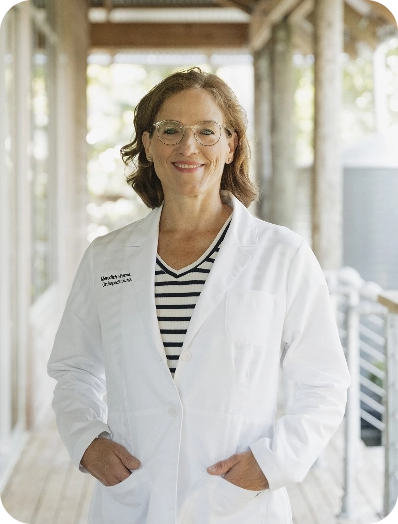 A smiling woman in glasses and a white lab coat stands indoors, representing surgeon-formulated supplements.